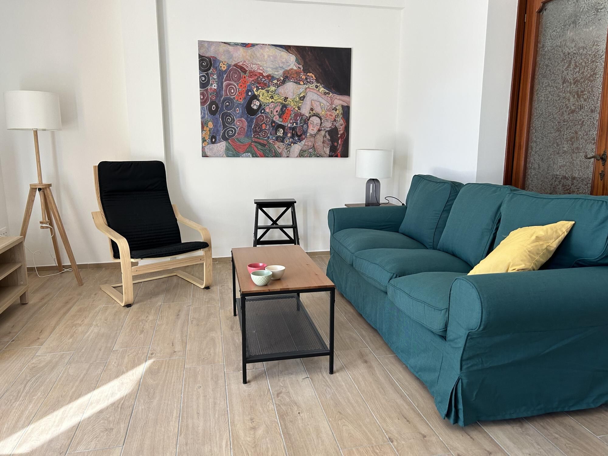 En stue med en tealgrøn sofa, en sort lænestol, et lille bord med skåle, en gulvlampe og et maleri på væggen.