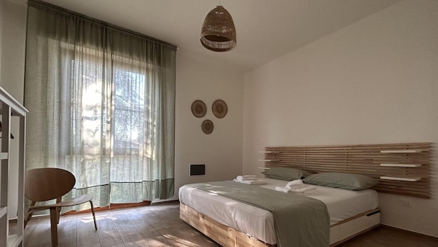 A bedroom with a wooden bed, a chair, a hanging light fixture, and a window with sheer curtains. - Casa La Pigna in Pisa (Pisa)