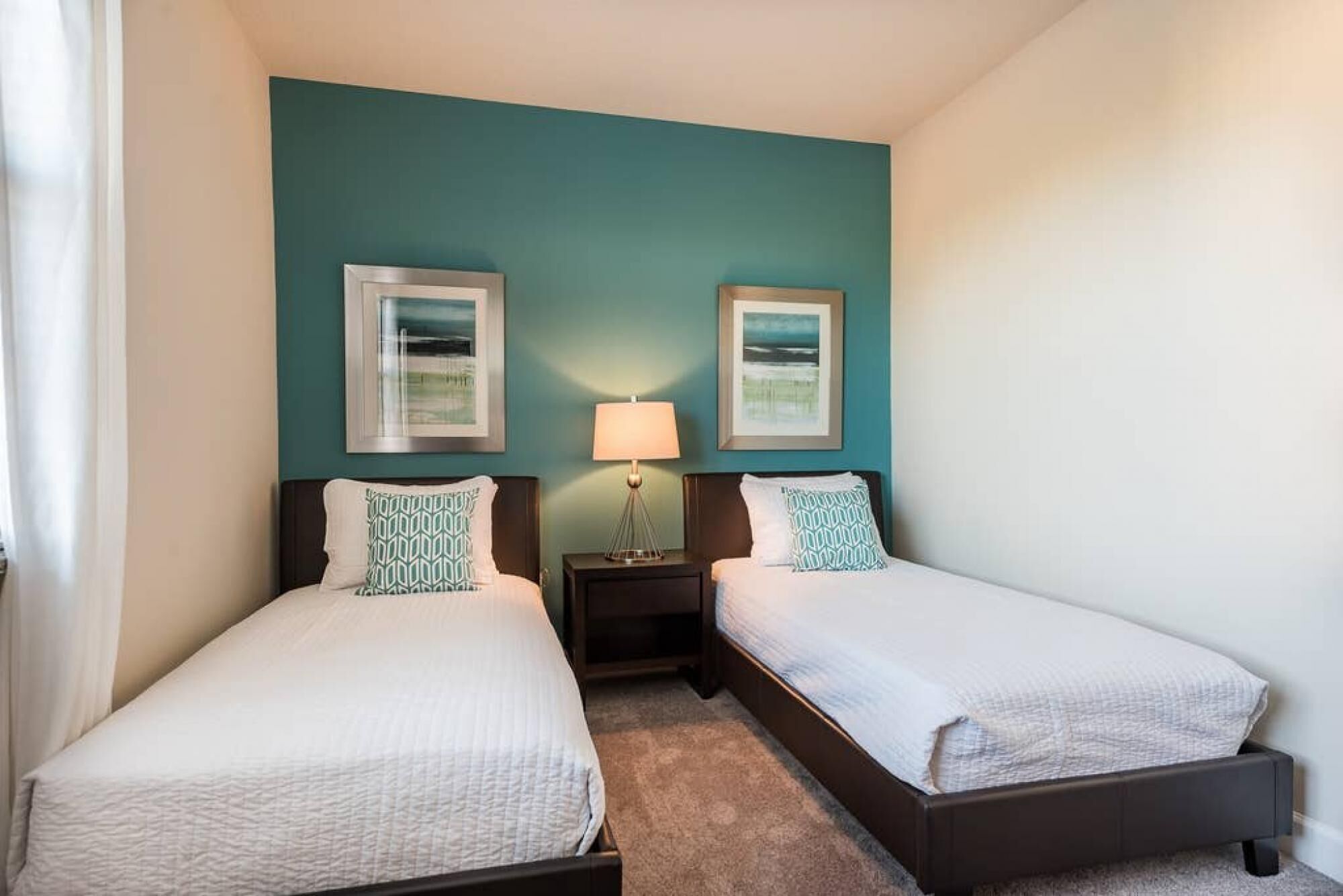 A hotel room with two single beds, a nightstand with a lamp, and two framed pictures on the wall.