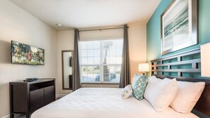 A hotel room with a large bed, a desk, a chair, a TV, and a window with blinds. - Super Cozy Home With Private Pool (Clermont)