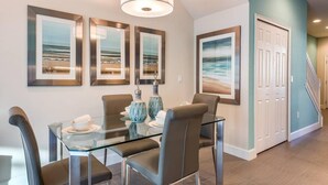 A dining area with a glass table, four chairs, and framed artwork on the wall. - Super Cozy Home With Private Pool (Clermont)