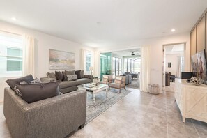 A spacious living room with a grey sofa, a coffee table, and a TV on a cabinet. There is a large window and a door leading to another room. - Stunning 7BR Retreat Private Pool Perfect Location (Kissimmee)