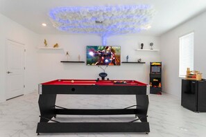 Uma mesa de bilhar em um quarto com luminária no teto, televisão e prateleiras com objetos decorativos.