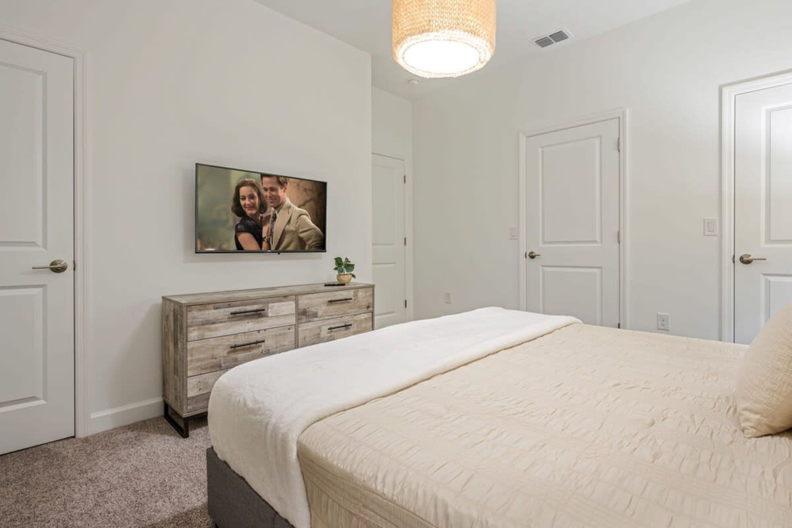 Une chambre avec un grand lit, une commode, un téléviseur fixé au mur et une porte sur le côté droit.