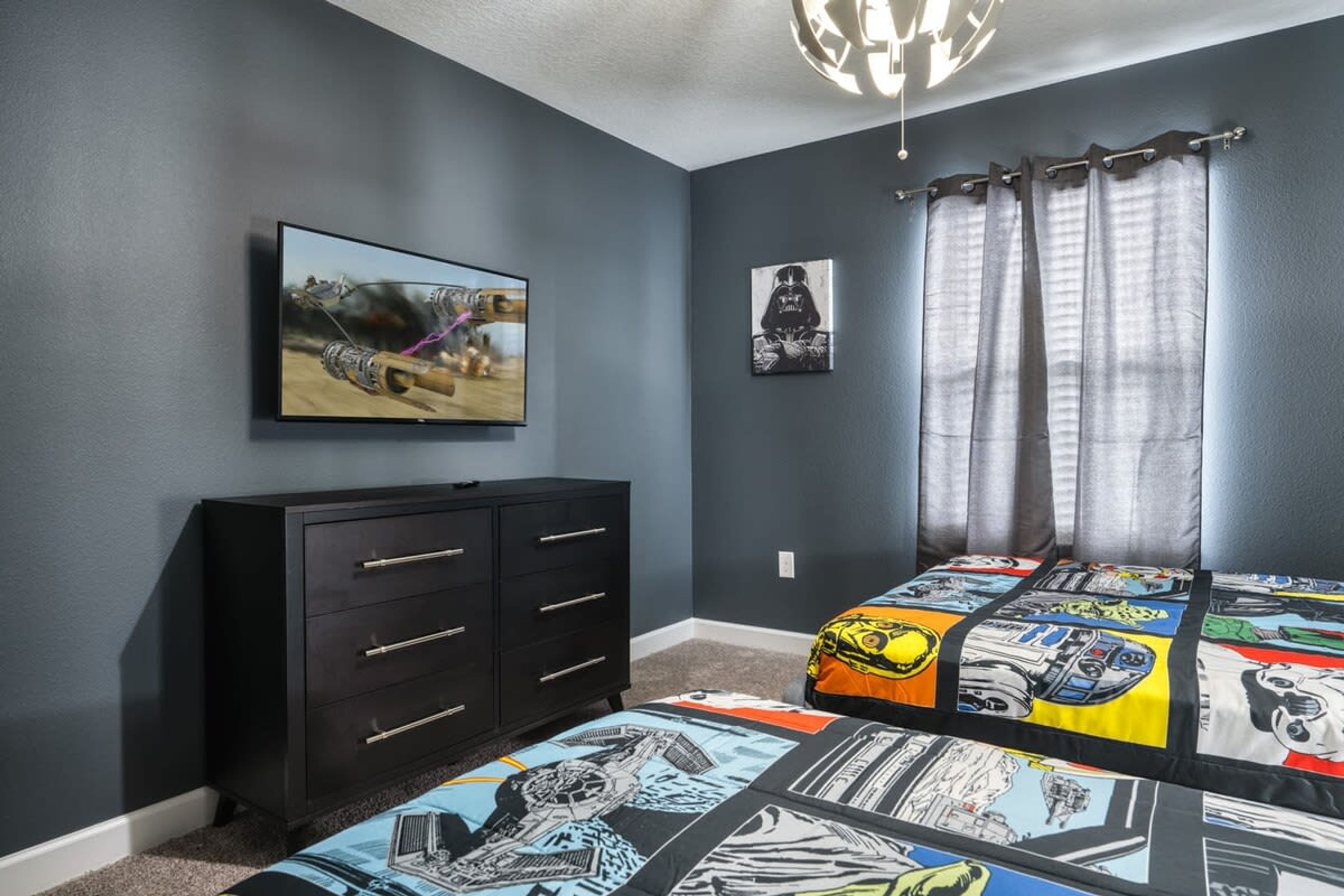 A bedroom with a dark blue wall, a black dresser with drawers, a bed with a comic book themed bedspread, a TV on the wall, and a Darth Vader poster.