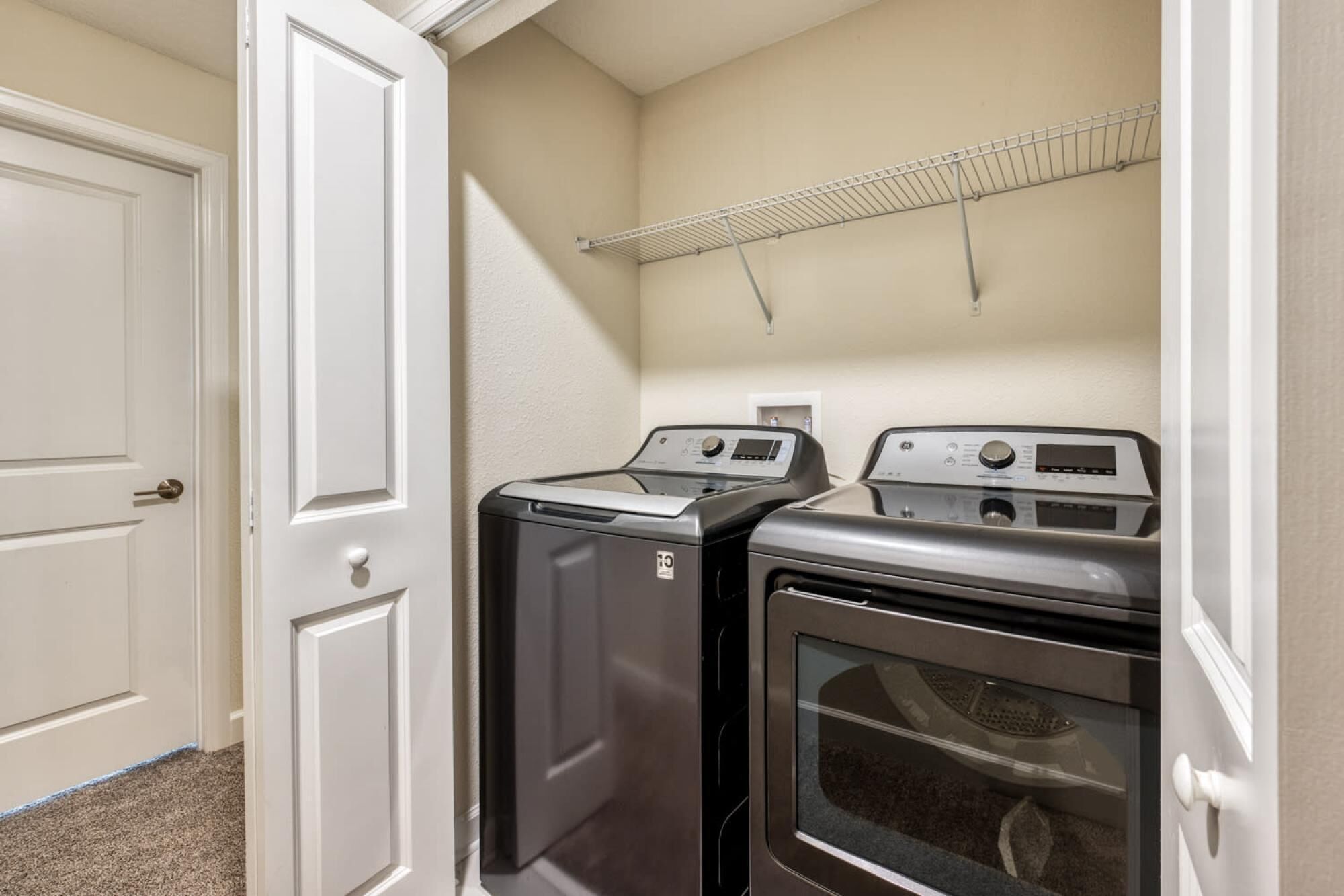 A laundry room with two washers and a dryer, a white door, and a metal drying rack on the wall.