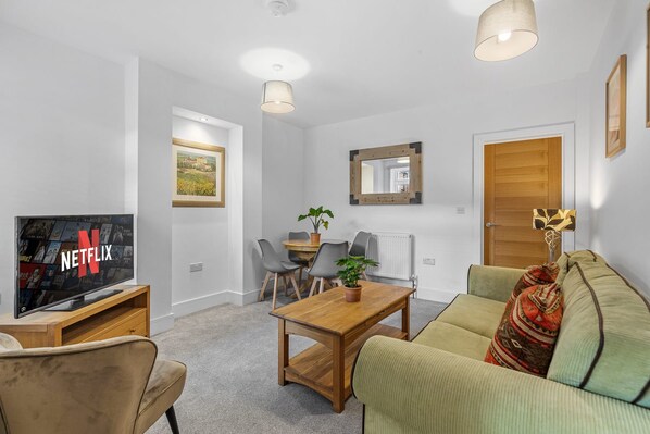 A modern living room with a flat-screen TV, a wooden coffee table, a sofa, and a dining area with chairs and a table.