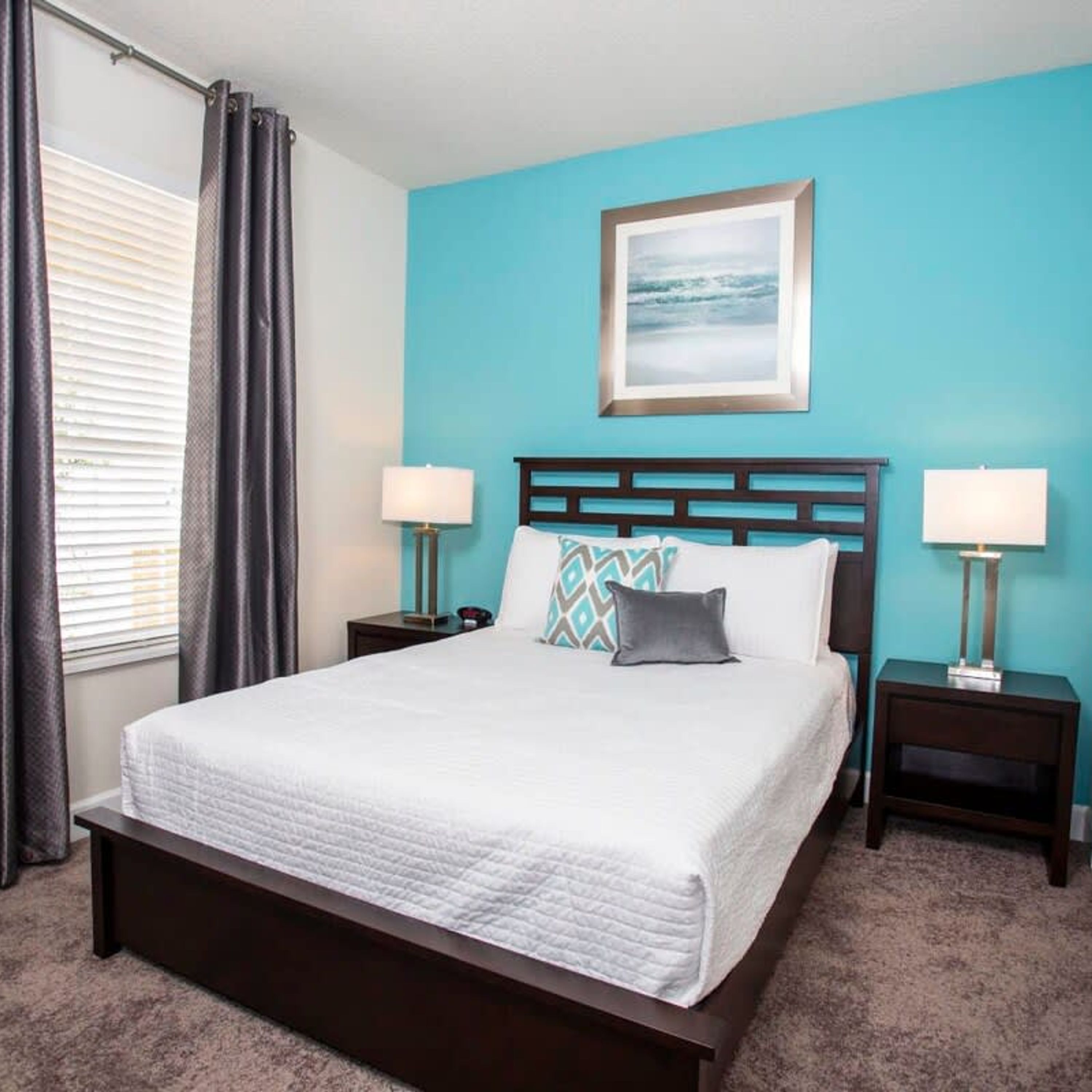 A neatly made bed with a white comforter and two pillows, flanked by two bedside tables with lamps, in a room with teal walls and a framed picture above the bed.