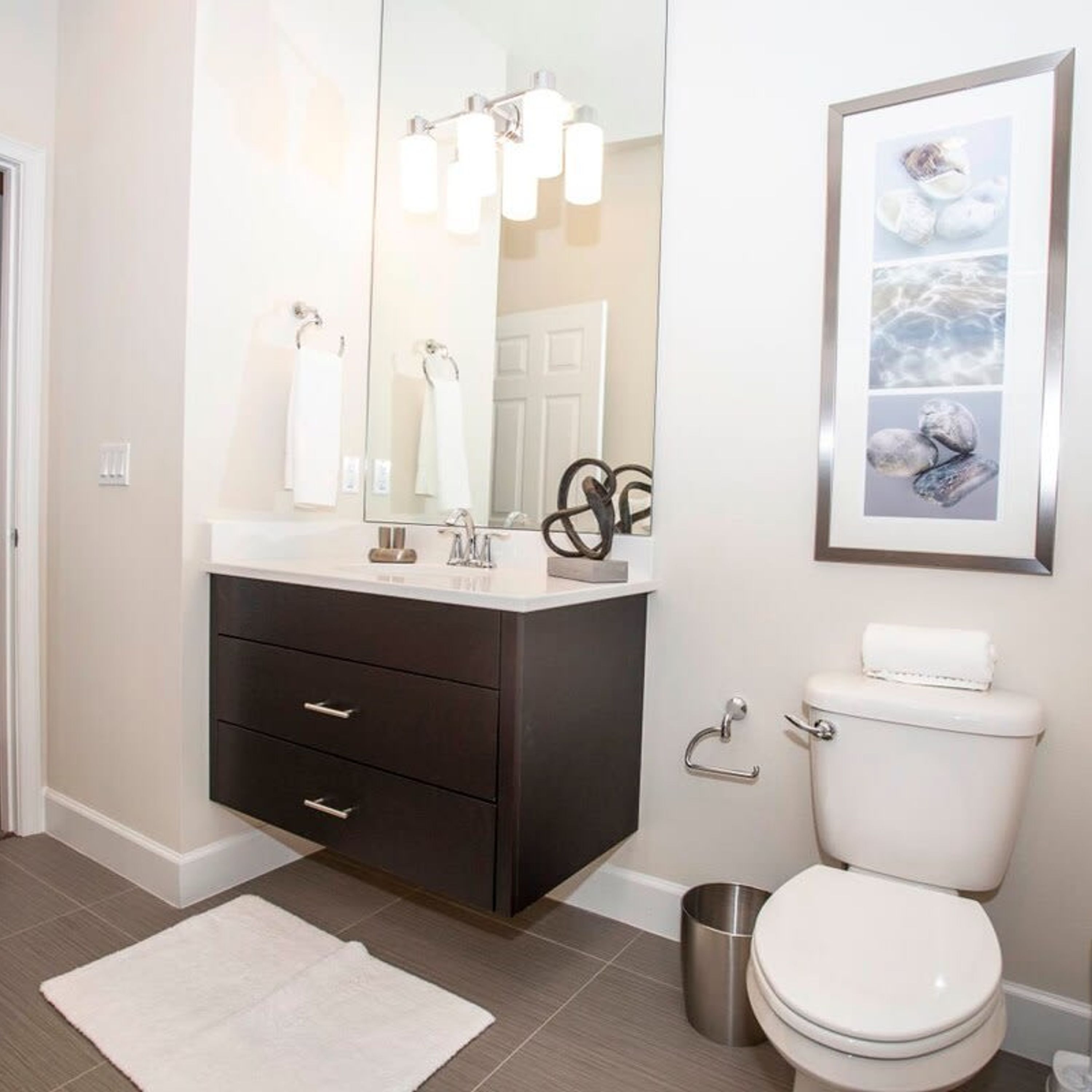 Une salle de bain moderne avec un meuble-lavabo foncé, un grand miroir, des toilettes blanches et une œuvre d’art encadrée au-dessus des toilettes.