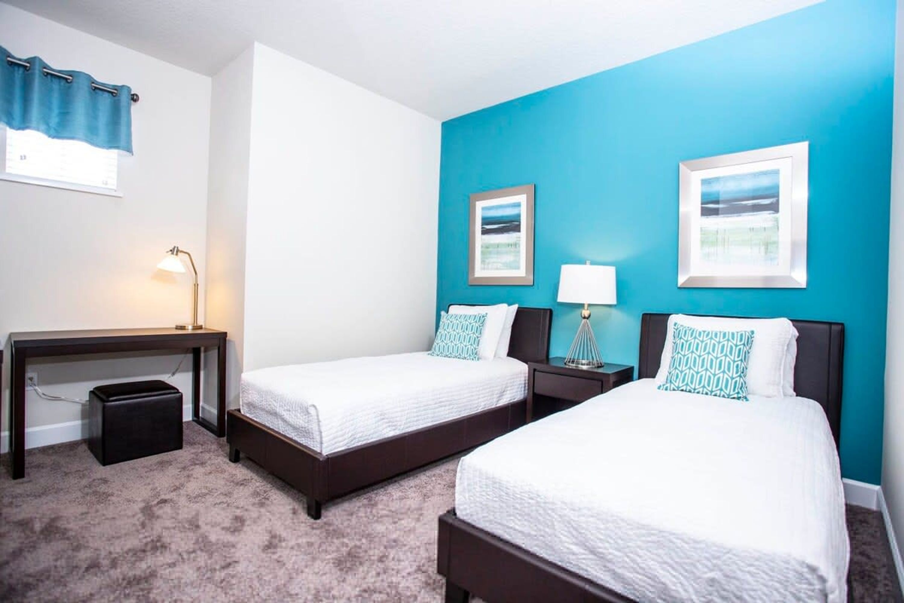 A hotel room with two beds, a desk, a lamp, and two framed pictures on the wall.