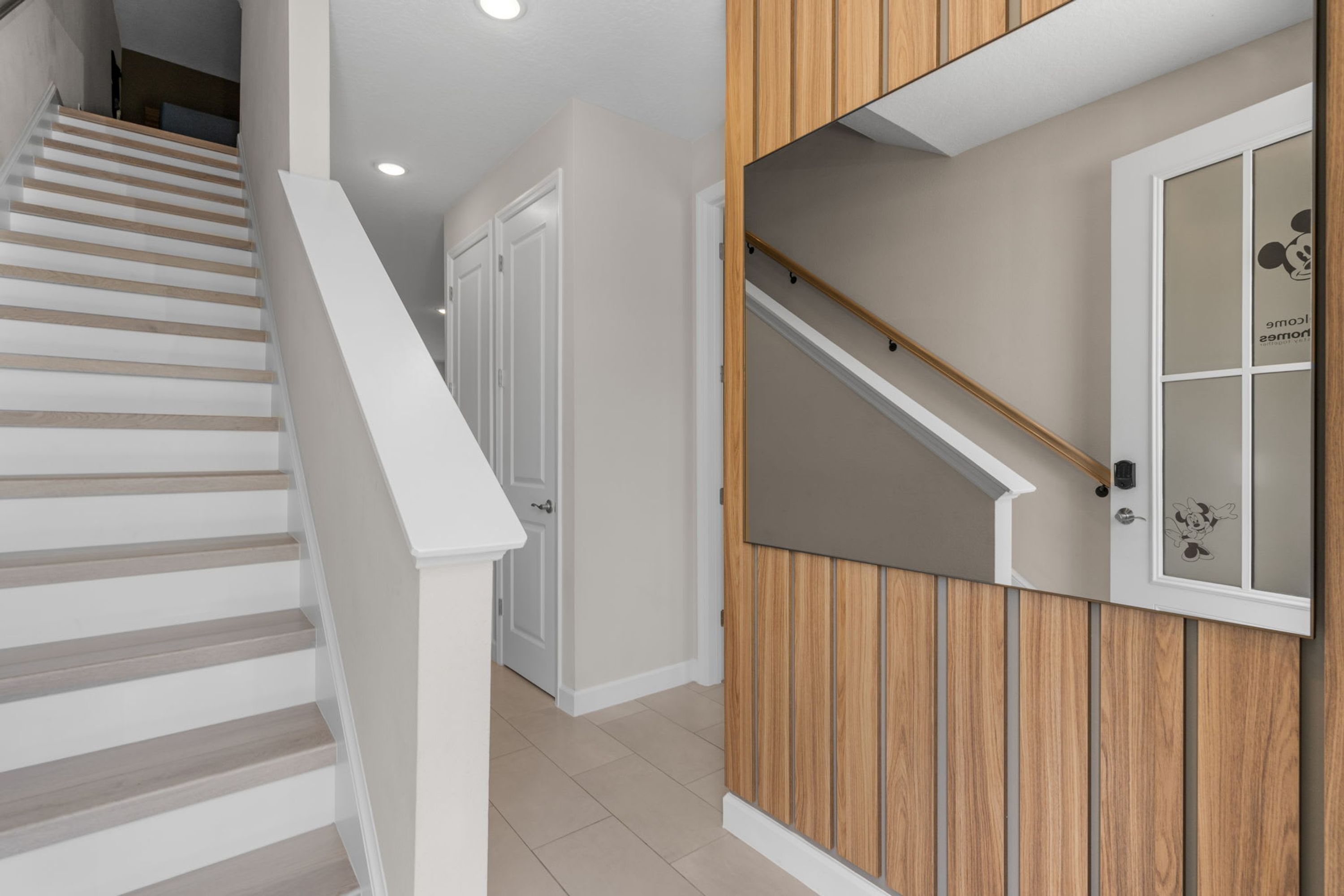 Un escalier avec des rampes blanches et des marches en bois, menant à une pièce dont un mur est recouvert de panneaux de bois et qui possède une porte ornée du logo de Mickey Mouse.