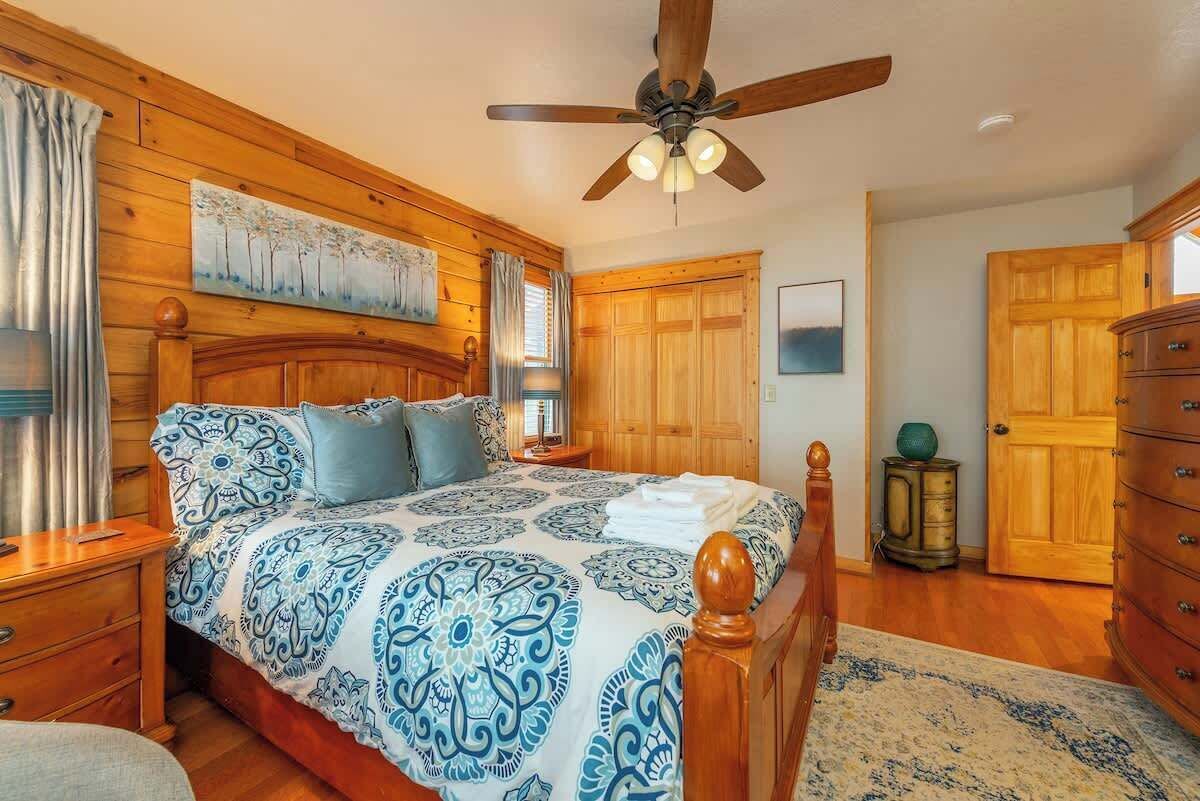 Un dormitorio con cabecera de madera, ventilador de techo, cama con ropa de cama estampada, mesita de noche, cómoda y puerta de madera.
