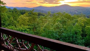 A balcony with a view of a forested landscape and a mountain range during sunset.