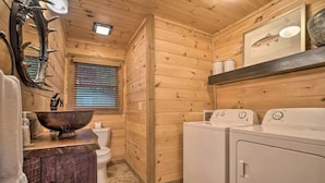 A wooden cabin interior with a wooden toilet, a large wooden bowl sink, and a set of white washing machines.