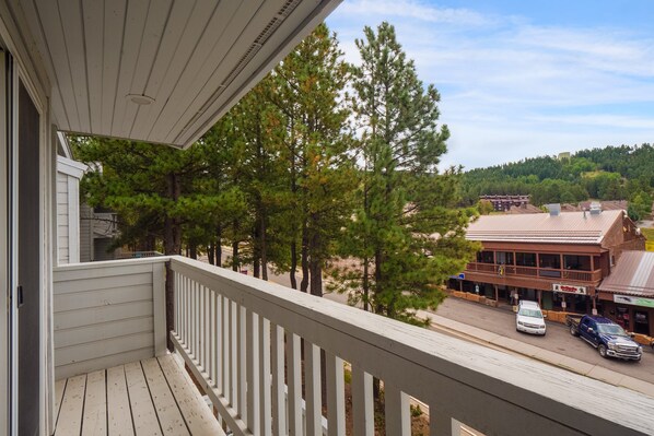 A wooden balcony with a view of a town street, buildings, and a forested hill.