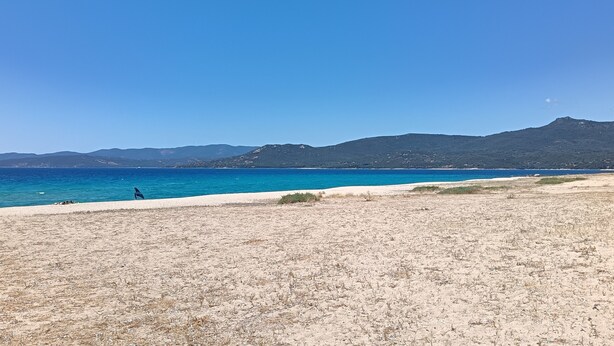 Una spiaggia con cielo sereno, montagne in lontananza e una persona che cammina sulla riva.