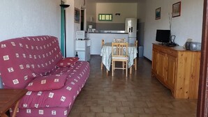 Un ambiente compatto con divano, tavolo da pranzo con sedie, angolo cottura e TV su un mobile in legno.