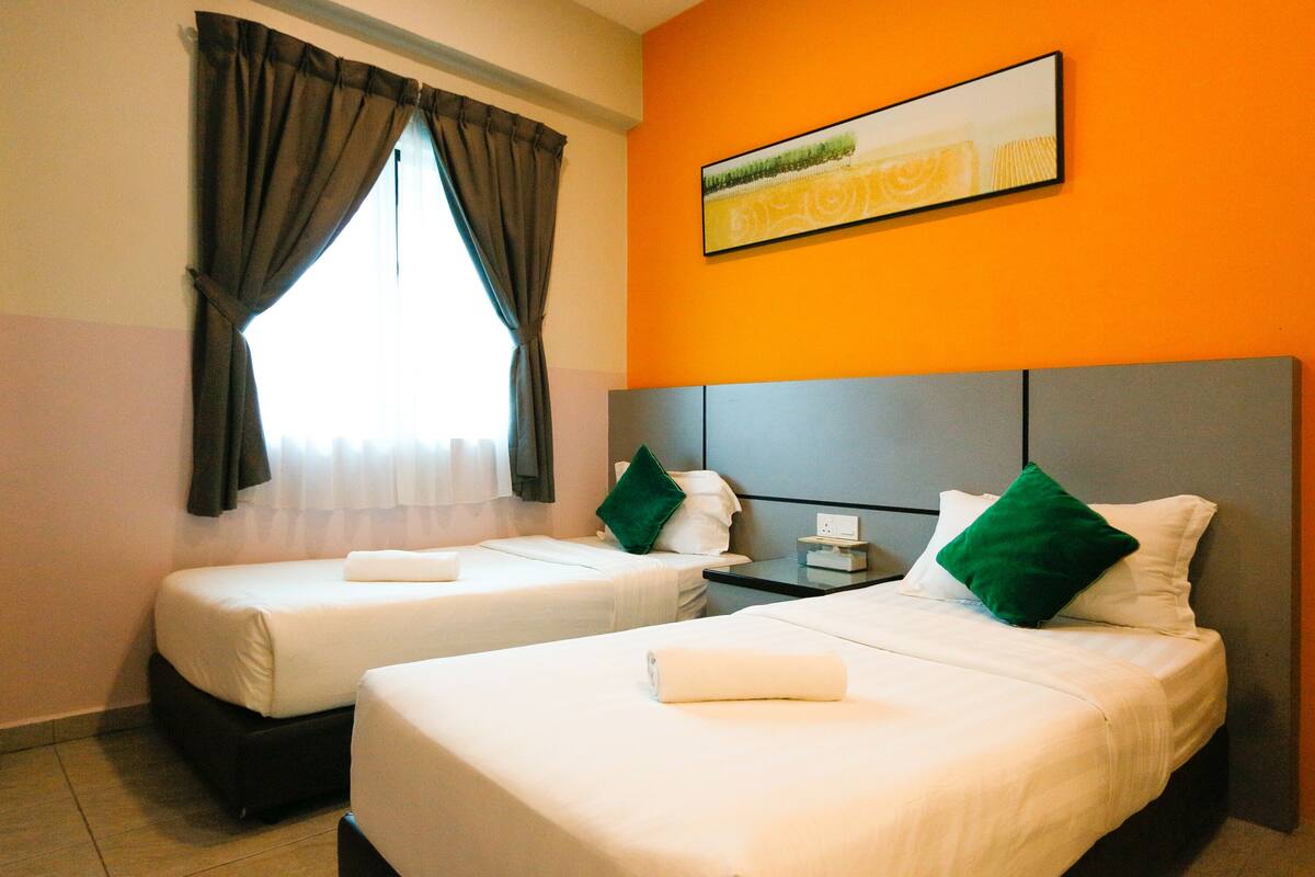 A hotel room with two beds, each with white linens and green pillows, a nightstand with a lamp, and a painting on the orange wall.