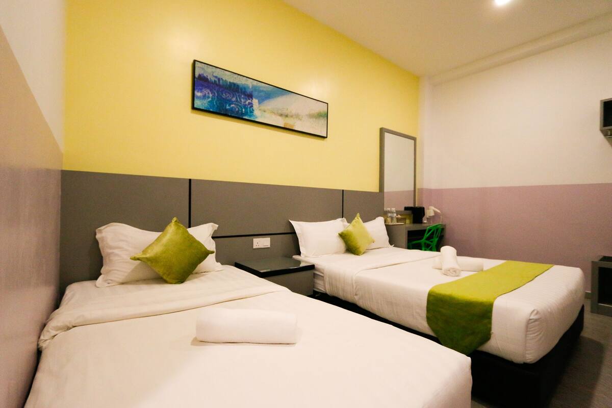 A hotel room with two beds, each with white bedding and green pillows, a small table, and a wall-mounted TV.