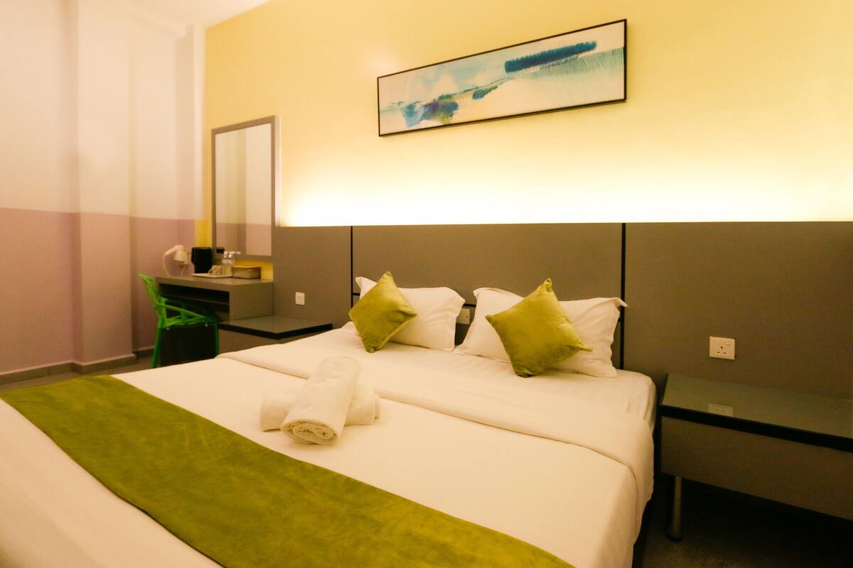 A hotel room with a large bed, green throw, white pillows, and a painting on the wall.
