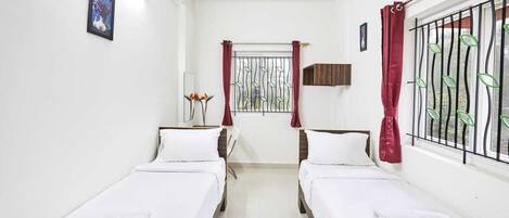 A room with two single beds, a window with metal bars, a small table with a lamp, and a picture on the wall.