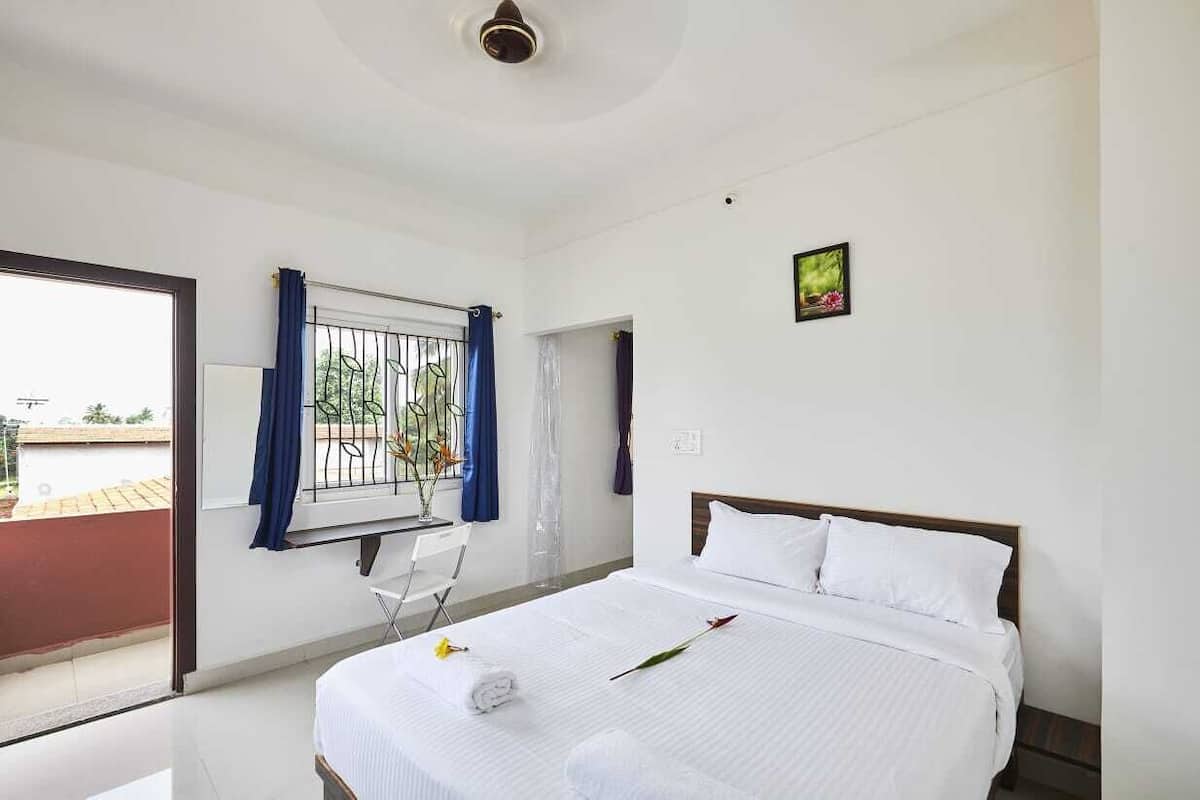 A hotel room with a bed, a window with blue curtains, a small table with a chair, and a view of the outside.
