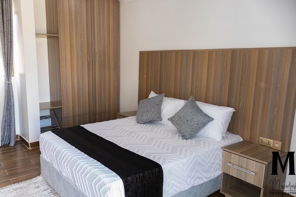 A neatly made bed with a white and gray patterned bedspread, two pillows, and a black throw blanket. There is a wooden nightstand with a drawer and a small table beside the bed. The room has wooden paneling on the walls and a window with sheer curtains.
