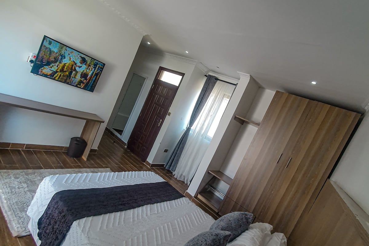 A bedroom with a bed, a wooden wardrobe, a painting on the wall, and a door leading to another room.