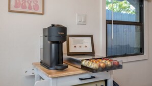 Eine Kaffeemaschine auf einem Wagen, mit einer Schale Kaffee-Kapseln, einem gerahmten Zertifikat und einem Fenster mit Jalousien.