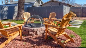 Ein Garten mit einem Steinkamin, umgeben von Holz-Adirondack-Stühlen, einer Holzterrasse und einem eingezäunten Bereich.