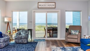 A spacious living room with a large sliding glass door leading to a deck with a view of the beach. There are two patterned armchairs, a blue sofa, and a small table with a lamp. - Sea La Vie (Port St. Joe)