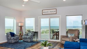 A spacious living area with a large window overlooking the beach, a ceiling fan, and a sign reading "BEACH HOUSE". - Sea La Vie (Port St. Joe)
