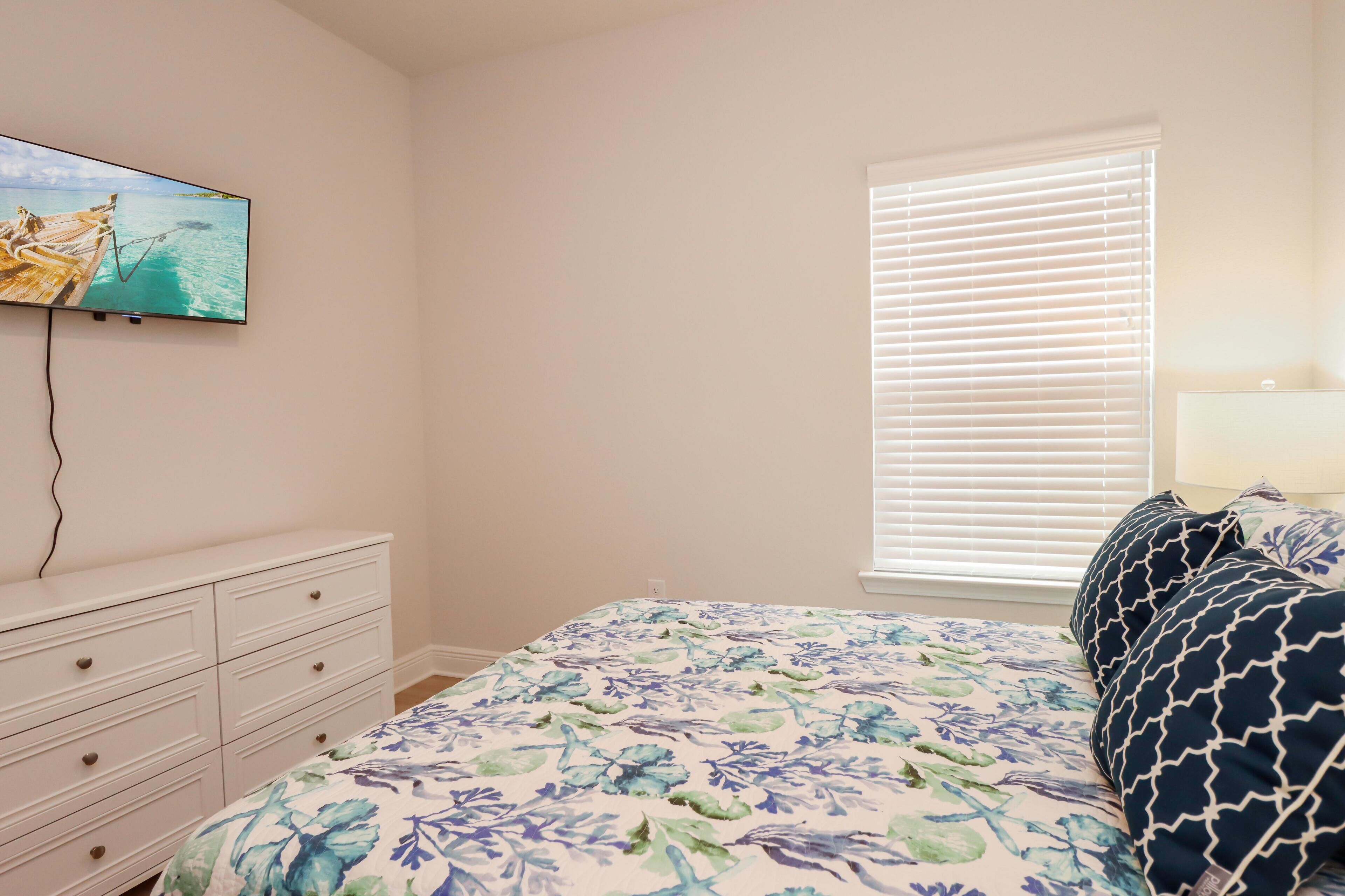Une chambre à coucher avec un lit, une commode, un téléviseur fixé au mur et une fenêtre avec des stores.