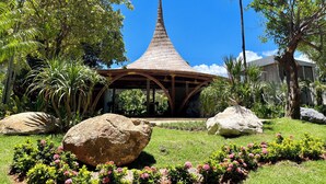 Un pavillon traditionnel doté d’un toit conique, entouré d’une végétation luxuriante et de gros rochers.