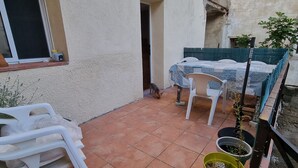 Un'area patio con pavimento piastrellato, una sedia di plastica bianca, un tavolo con tovaglia blu e bianca e una pianta in vaso.