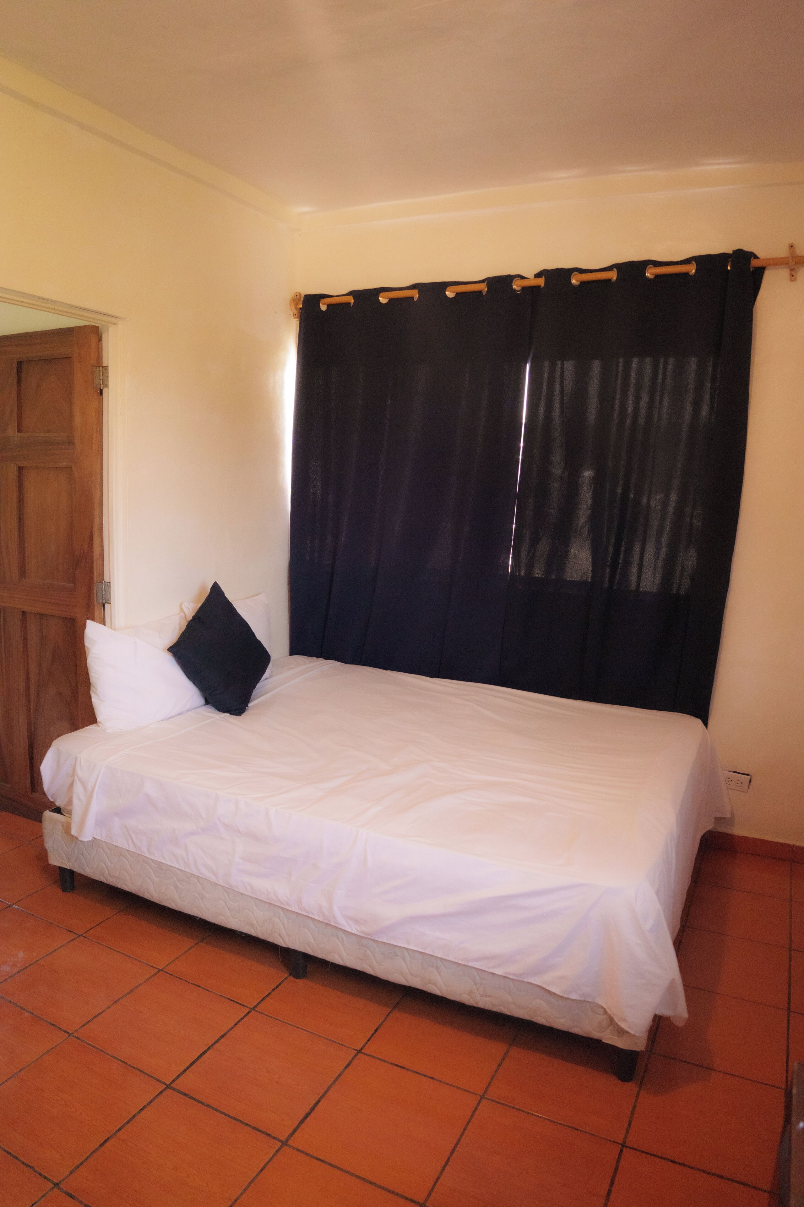 A single bed with a white mattress and a dark headboard, a wooden door to the left, and a tiled floor.