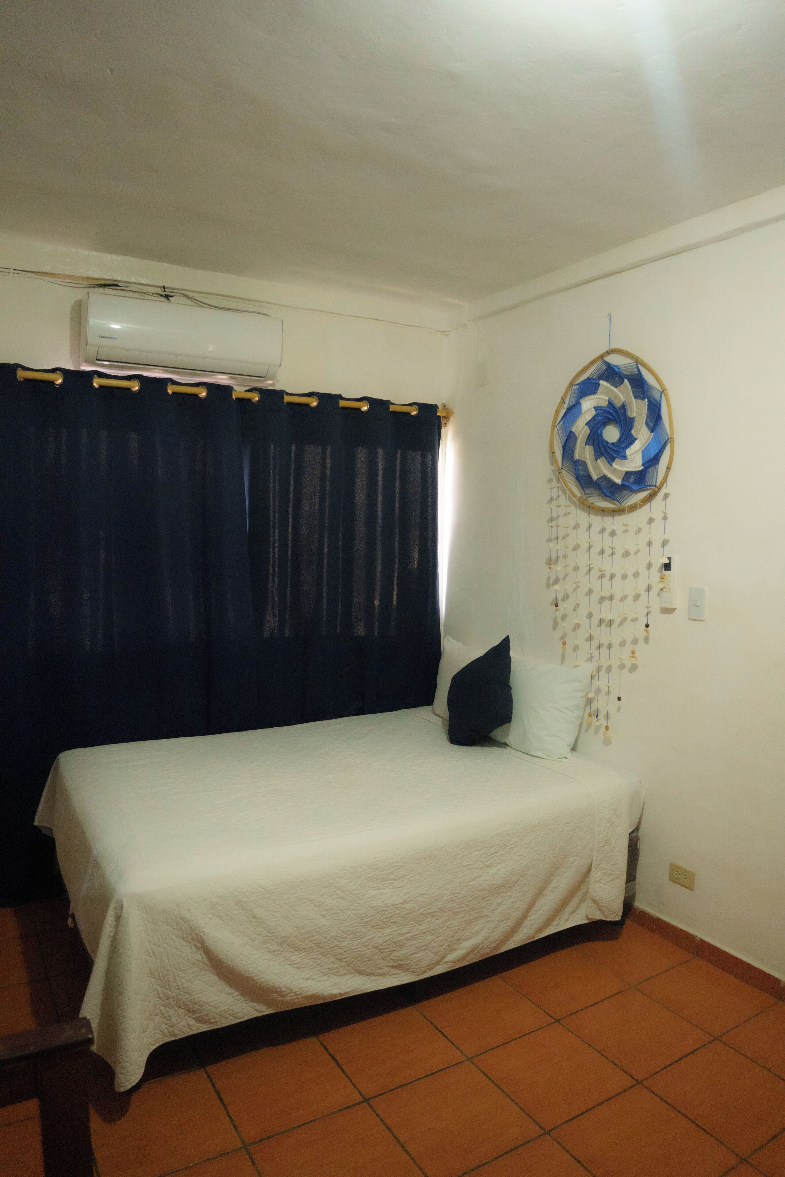 A bedroom with a bed, a wall-mounted air conditioner, a curtain, and a dreamcatcher.