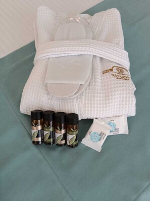 A neatly folded white robe with a logo, four bottles of olive care shower products, and a bar soap packet on a teal surface. - Navarino Luxe Suites with Sea View (Thermaikos)