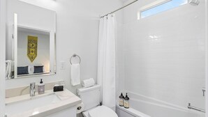 A modern bathroom with a white sink, toilet, and bathtub. There is a shower curtain, a towel rack, and a window providing natural light. - Stag Sweet Suite in Capitol Hill (Seattle)