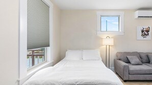 Une chambre avec un lit, une fenêtre avec des stores, une lampe de chevet, un canapé gris et une petite fenêtre au-dessus du lit.