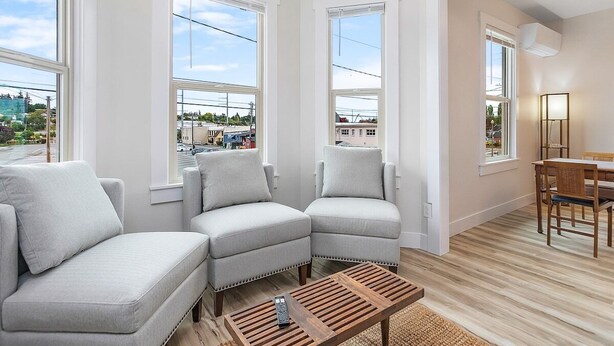 A modern living room with a sectional sofa, a coffee table, and a dining area with a table and chairs. Large windows provide natural light and views of the outside. - The Night Owl Perch in Stanwood (Stanwood)