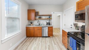 A compact kitchen with wooden cabinets, a dishwasher, a stove, and a countertop with various appliances and utensils. - The Night Owl Perch in Stanwood (Stanwood)