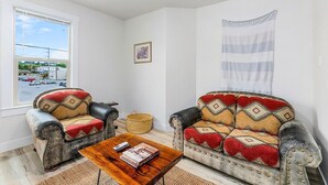 A living room with two patterned sofas, a wooden coffee table, a framed picture, and a view of a parking lot through a window. - The Nightjar Shelter (Stanwood)