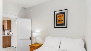 A hotel room with a bed, a nightstand, a lamp, a framed sign on the wall, and a door leading to a kitchen area. - The Nightjar Shelter (Stanwood)