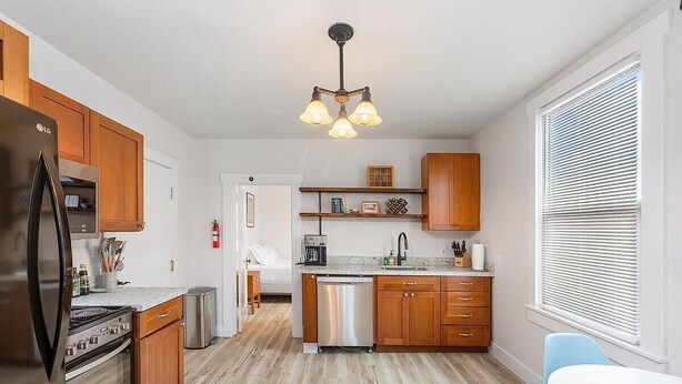 A modern kitchen with wooden cabinets, a stainless steel refrigerator, and a dishwasher. There is a dining area with a white table and blue chairs. - The Nightjar Shelter (Stanwood)