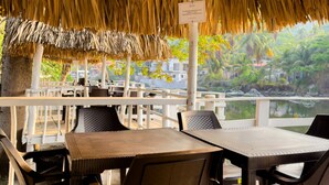 An outdoor seating area with thatched roof, wooden tables, and chairs, overlooking a body of water and greenery. - Playa Linda Surf Hotel (Tamanique)