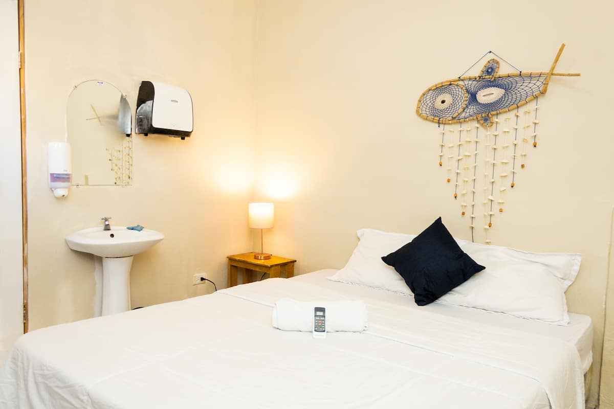 Hotellihuone, jossa on sänky, yöpöytä lampun kanssa, seinään kiinnitetty lämmitin, peili, pesuallas ja koristeellinen seinäkoriste.
