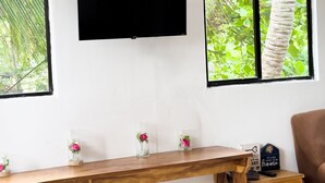 A room with a wooden desk, a mounted TV, and two windows with a view of greenery. - Playa Linda Surf Hotel (Tamanique)