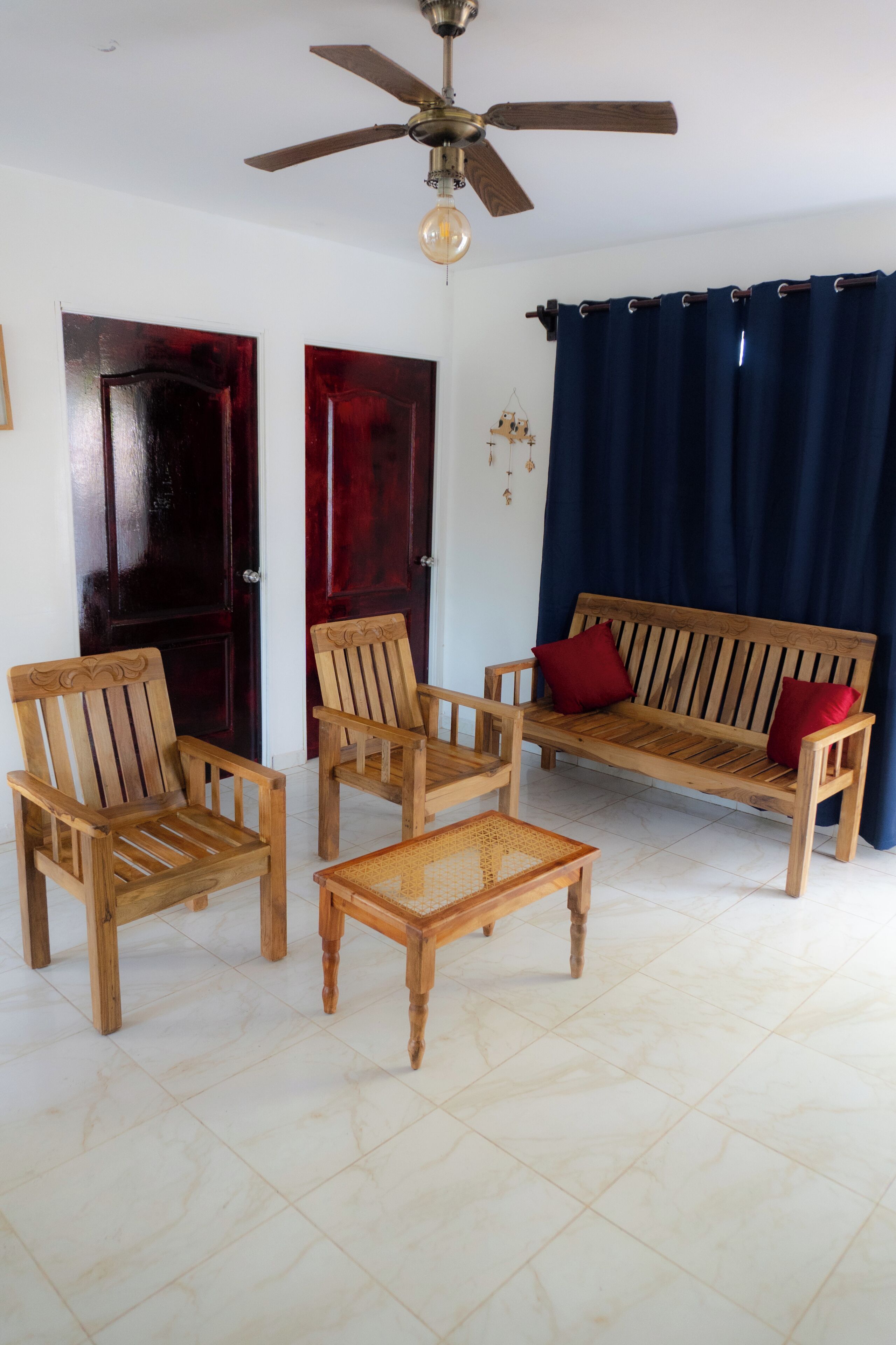 Una sala de estar con ventilador de techo, un sofá de madera con cojines rojos, dos sillas de madera y una pequeña mesa de madera.