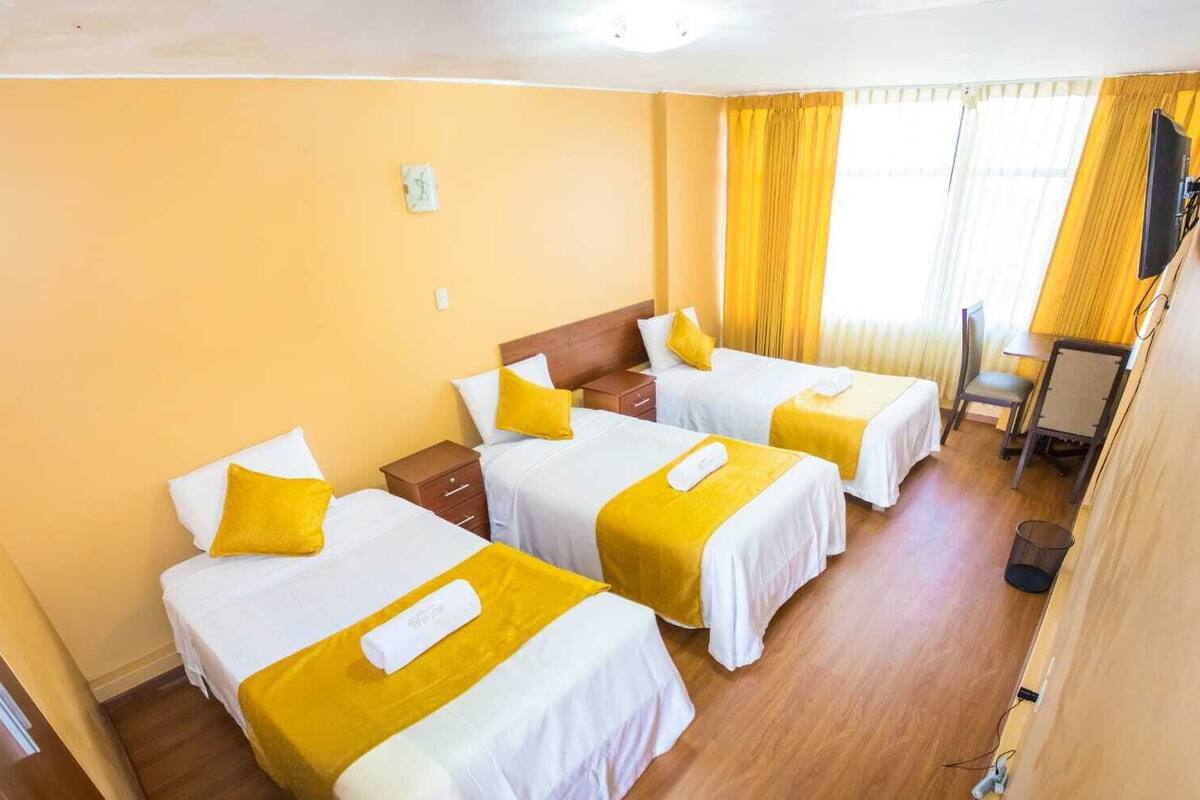 Habitación de hotel con cuatro camas, cada una con ropa de cama blanca y detalles amarillos, suelo de madera, un escritorio pequeño y un televisor montado en la pared.